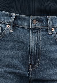 Κοντινή λήψη της μέσης παντελονιού από μπλε τζιν με κούμπωμα κουμπιού και μέρος μαύρου ριπ πλεκτού που έχει μπαίνει μέσα.