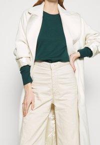 Kvinna iklädd en vit lång kappa över en mörkgrön tröja och högmidjade benvida byxor i off-white, med handen på höften och avslappnad hållning.