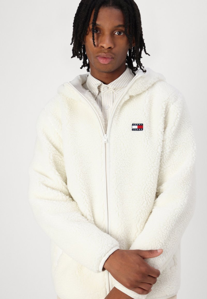 Sweat à capuche zippé blanc en polaire, avec une surface texturée, arborant un patch logo sur la poitrine et une capuche doublée. Le modèle se tient les bras croisés.