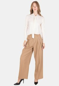 Blusa color crema con collo a fiocco, abbinata a pantaloni beige a gamba larga e vita alta e tacchi a spillo neri. Tessuto liscio, vestibilità comoda.