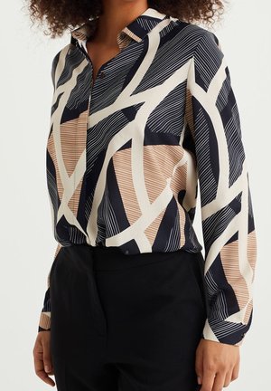 Vrouw draagt een blouse met lange mouwen en knopen, met abstracte geometrische patronen in marineblauw, beige en crème, ingestopt in een zwart broek met een hoge taille.