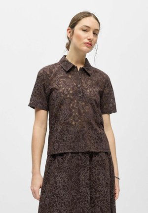 Femme portant une chemise marron à manches courtes boutonnée et une jupe assortie avec des motifs floraux et feuillus complexes.