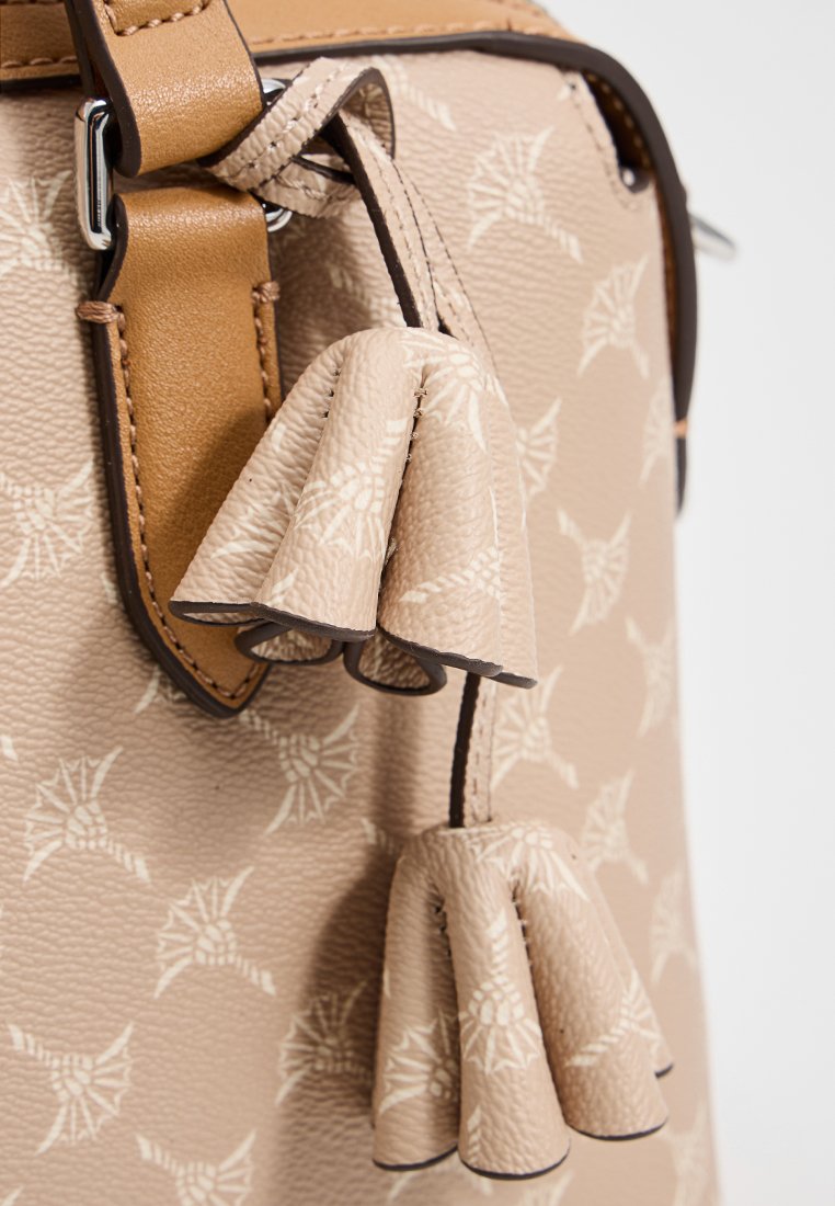 Beige strukturiert Tasche mit gemusterter Oberfläche, ausgestattet mit braunen Lederakzenten und zwei dekorativen Quasten. Silberne Beschläge sind sichtbar.