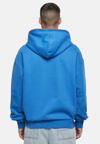 Niebieska bluza z kapturem, wykonana z mieszanki bawełny, wyposażona w ściągacze na mankietach i dole, luźny krój oraz duży kaptur o bezszwowej konstrukcji.