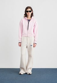 Γυναίκα που στέκεται φορώντας μαύρα γυαλιά ηλίου, ροζ φούτερ με φερμουάρ, κρέμα παντελόνια φόρμας με λογότυπο και λευκά αθλητικά παπούτσια σε μπλε χαλί.