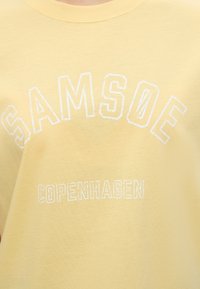 Κίτρινο ύφασμα με λευκό κείμενο «SAMSOE» σε τόξο πάνω από το «COPENHAGEN» με casual, ελαφρώς φθαρμένη γραμματοσειρά.