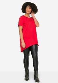 Tunic top asimmetrica rossa con maniche corte, abbinata a leggings in pelle nera e stivaletti con borchie, con una texture liscia.