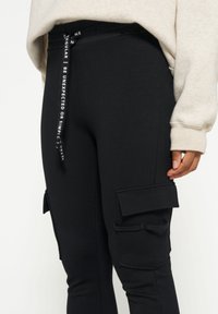 Zwarte cargobroek met een elastische tailleband, zijzakken met kleppen en een trekkoord met tekst. Zacht stof met een gladde textuur.