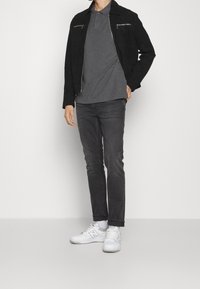 Veste en daim noire avec poches zippées, polo gris, jean gris foncé et baskets blanches. Design simple sans motifs visibles.