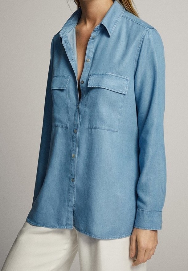 Femme portant une chemise en denim bleu clair boutonnée avec des poches poitrine et un pantalon blanc sur un fond uni.