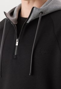 Sudadera negra con cremallera y capucha forrada en gris, tejido texturizado, cremallera metálica y un discreto logo bordado en el pecho.