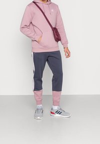 Růžová mikina s kapucí a klokáním kapsou s logem, doplněná tmavě modrými tepláky s růžovými manžetami a bílými bočními pruhy. Šedé sportovní boty a crossbody taška.