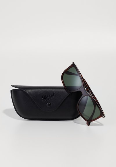 Óculos de sol em padrão tartaruga castanha com lentes verde-escuro, repousando ao lado de uma caixa de couro preta com fecho de pressão e logotipo em relevo.