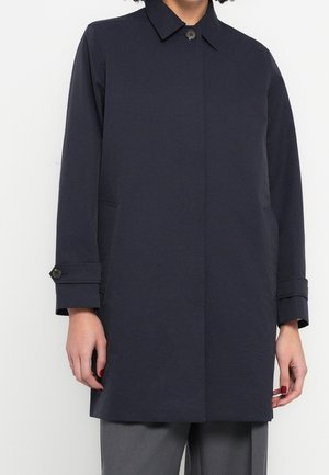 Vrouw draagt een donkerblauwe jas met een enkele knoop, zijzakken en knoopmanchetten over een grijze broek, staand tegen een effen achtergrond.