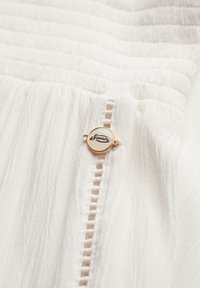 Prenda de tela blanca con pliegues texturizados, que presenta un acento de logotipo circular en tono dorado con texto negro. Se muestra un delicado detalle de costura.