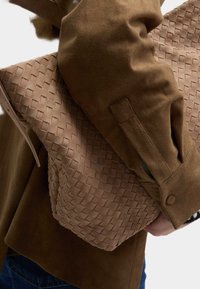 Sac en cuir tressé marron tenu contre un vêtement en tissu marron texturé, présentant des matériaux lisses et doux avec un motif géométrique sur le sac.