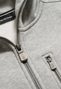Primer plano de una chaqueta de tela gris claro con dos cremalleras de metal plateado, una para la abertura principal y otra más pequeña para un bolsillo.