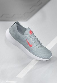 Svijetlosivi tenisice s teksturiranim mrežastim gornjim dijelom, bijelim gumenim potplatom i istaknutim crvenim Nike logom sa strane i na jeziku.