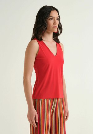 Donna con capelli scuri ondulati che indossa una maglia rossa senza maniche con scollo a V e pantaloni a righe verticali multicolore, in piedi davanti a uno sfondo chiaro e semplice.