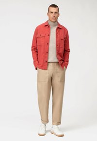 Rote Jacke mit Knöpfen und zwei Brusttaschen, beigefarbene Hose mit Seitentaschen, weiße Sneakers. Hellgrauer Rollkragenpullover darunter.