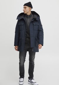 Marineblå pufferjakke med hætte, der har trykknaplommer, lynlås og en glat stoffinish. Bæres over en grå top og sorte jeans.