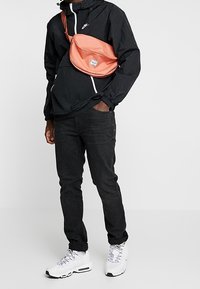 Person klädd i svart Nike-jacka, svarta jeans, vita sneakers och en korallfärgad axelväska mot en plain vit bakgrund.