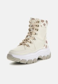 Botas altas de color crema con un collar acolchado, ojales dorados y una suela texturizada en blanco y gris. Presenta un patrón de logo marrón en la tela.