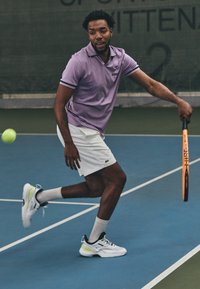 Vyras, vilkintis levandų spalvos polo marškinėlius ir balta šortus, ruošiasi smūgiuoti teniso kamuolį, laikydamas oranžinį teniso raketę mėlyname korte.