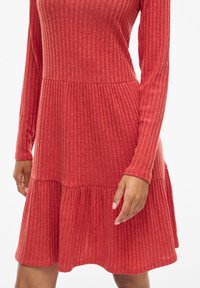 Červené žebrované šaty s dlouhými rukávy, plisovanou sukní a texturovanou tkaninou. Zahrnují upnutou korzetovou část a rozšířený lem.