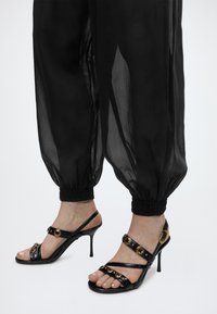 Πόδια που φορούν μαύρα σανδάλια με ψηλό τακούνι, διακοσμημένα με χρυσά καρφιά και αγκράφες, συνδυασμένα με διάφανη μαύρη παντελόνα με σούρα στους αστραγάλους.