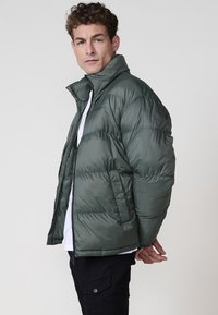 Chaqueta verde acolchada con cuello alto, cierre de cremallera y bolsillos laterales. Textura suave con un diseño acolchado.