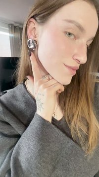 Brincos de prata com formas abstratas numa pessoa de cabelo comprido, vestindo um suéter cinzento. Destacam-se o design geométrico e o acabamento brilhante.