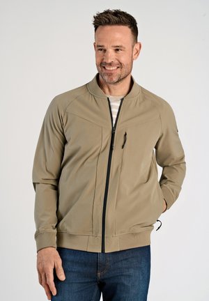 Giacca bomber beige con zip frontale, due tasche sul petto e polsini a coste, abbinata a jeans blu scuro e una camicia a righe sotto.