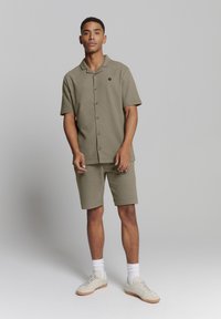 Korte mouw khaki knopshirt gecombineerd met bijpassende shorts. De stof heeft een zachte textuur, met een subtiele merkaankleding op het shirt.