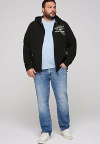 Chaqueta negra con cremallera y capucha, con gráficos en blanco. Camiseta de cuello en V azul claro debajo. Jeans azul claro y zapatillas blancas.