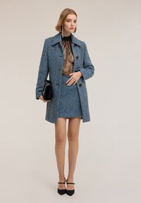 Cappotto in tweed blu e nero con bottoni grandi, indossato sopra una maglietta nera trasparente. Abbinato a una gonna coordinata e una borsa nera, con tacchi a punta.