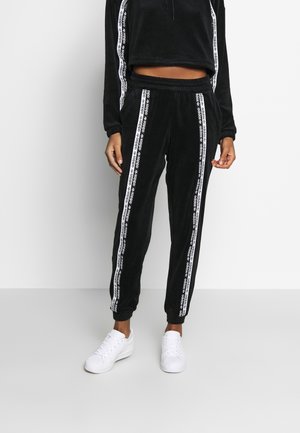 Personne portant un pantalon de survêtement Adidas noir et un sweat à capuche court assorti avec des bandes blanches au logo, associée à des baskets blanches, debout contre un mur blanc.