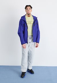 Μωβ ελαφρύ μπουφάν με μαύρες λεπτομέρειες, κοντομάνικο πράσινο μπλουζάκι, ανοιχτό μπλε τζιν και μαύρα αθλητικά παπούτσια με μπλε σόλες.