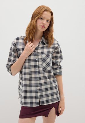 Giovane donna con capelli rossi che indossa una camicia a quadri beige e nera con le maniche arrotolate e una gonna bordeaux con uno spacco frontale su uno sfondo semplice.