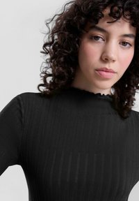 Zwarte geribbelde top met een hoge halslijn en een ruffled rand. Heeft een aansluitend ontwerp met textuurstof en lange mouwen.