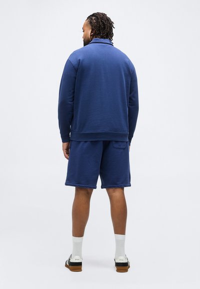 Άνδρας με πλεξούδες που φοράει μπλε σκούρο φούτερ και ταιριαστά σορτς, άσπρες κάλτσες και ασπρόμαυρα αθλητικά παπούτσια, στέκεται με την πλάτη γυρισμένη.