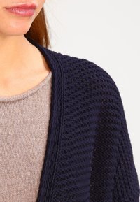 Marinblå stickad cardigan med öppen fram, med en texturerad väv, bärs över en ljusbeige topp. Närbild av krageområdet visas.