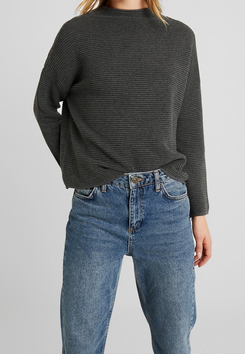 Dunkelgrauer, langärmeliger, gerippter Pullover mit hohem Kragen, getragen über hellblauen, hochtaillierten Jeans. Der Stoff hat eine strukturierte Optik.