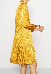 Žluté šaty z měkké, lesklé tkaniny s tečkovanou texturou. Mají dlouhé rukávy a rozšířený, volánkový lem.