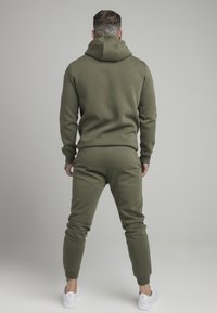 Olive green hoodie and joggers set, made of soft fabric. Features a hood, ribbed cuffs, and tapered legs, paired with white sneakers.