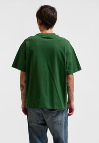 Green cotton t-shirt with short sleeves, relaxed fit, and crew neckline. Visible stitching and textured fabric, paired with light blue jeans.