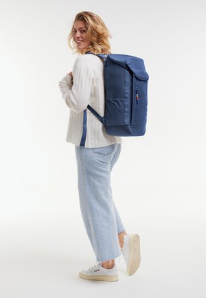 Nainen hymyilee olkansa yli valkoinen neule yllään, vaaleansiniset farkut, valkoiset lenkkarit ja suuri sininen reppu selässään.