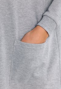 Ljusgrå sweatshirt med en stor framficka, ribbade ärmslut och en mjuk textur. Fokusera på fickans detalj och tygkomposition.