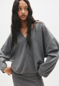 Grå oversized sweater med en dyb V-udskæring og lange, brede ærmer. Stoffet er blødt med en glat overflade, der komplimenterer en tætsiddende grå nederdel.