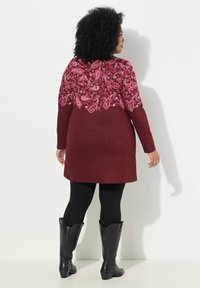 Dlouhé vínové šaty s růžovým paisley vzorem na horní části zad, dlouhé rukávy, nošené přes černé legíny, doplněné černými kovbojskými botami.